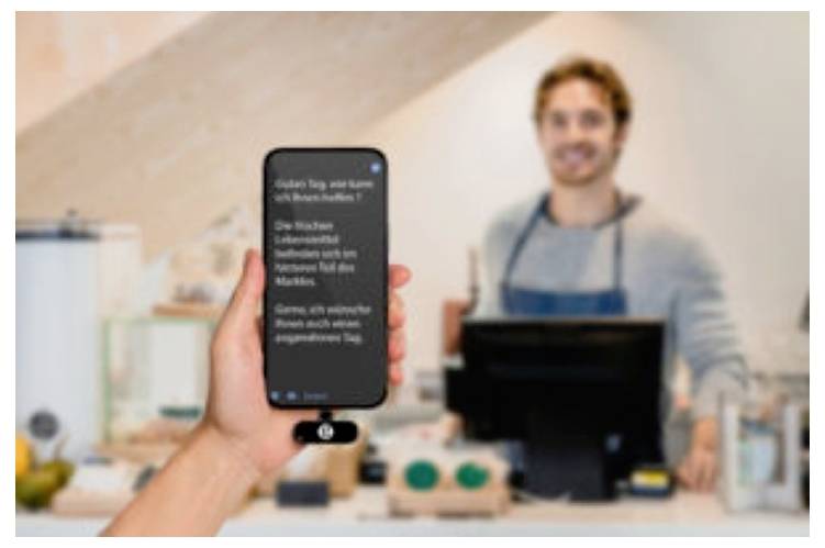 Hand hält ein Smartphone mit Text zu einem lächelnden Café-Kassierer hinter der Theke, der eine Mobile-Payment- oder Bestellungs-App zeigt.
