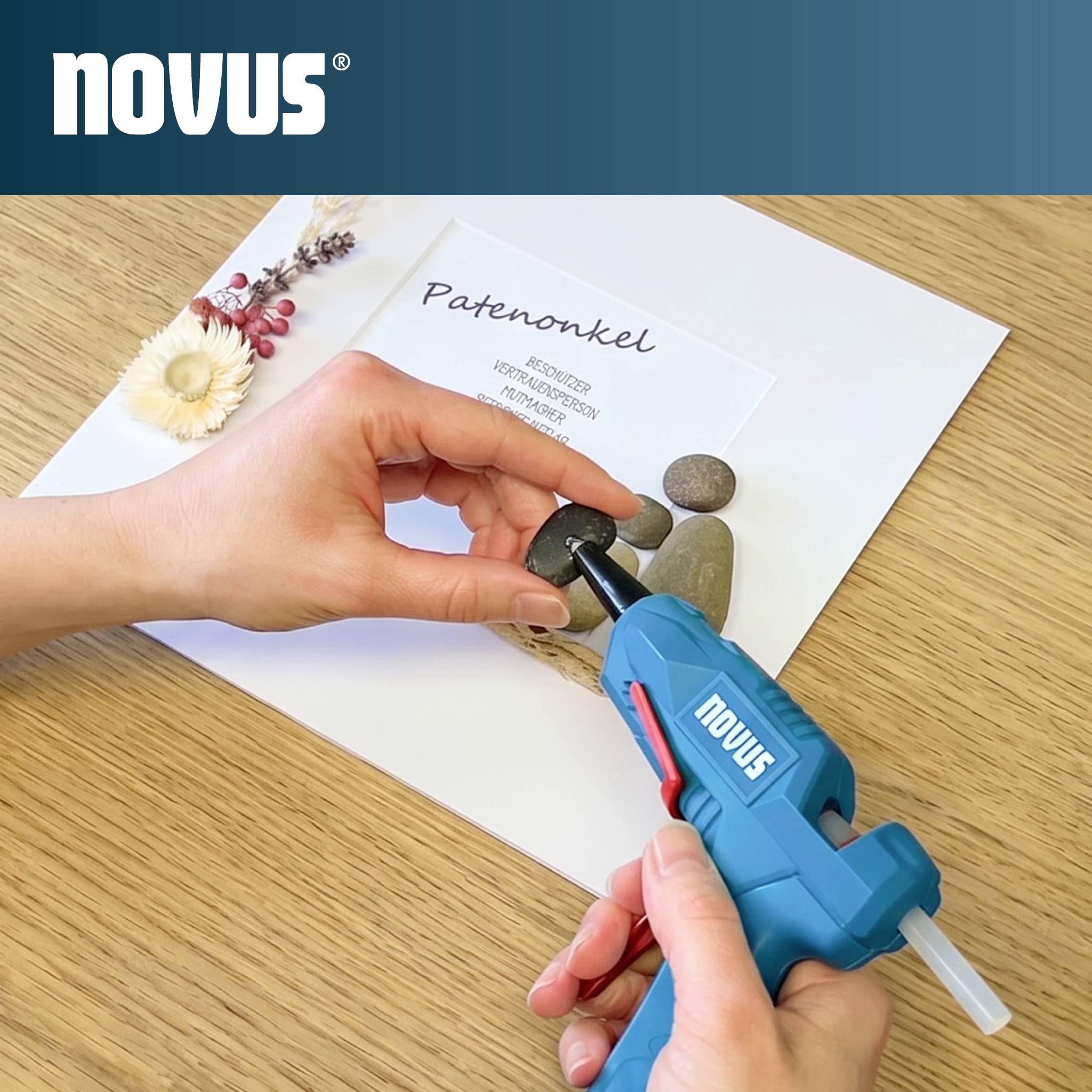 Hände, die eine blaue Novus-Heißklebepistole verwenden, um einen kleinen Stein an ein Papierkraftprojekt mit der Bezeichnung "Patenkel" auf einem Holztisch anzubringen.
