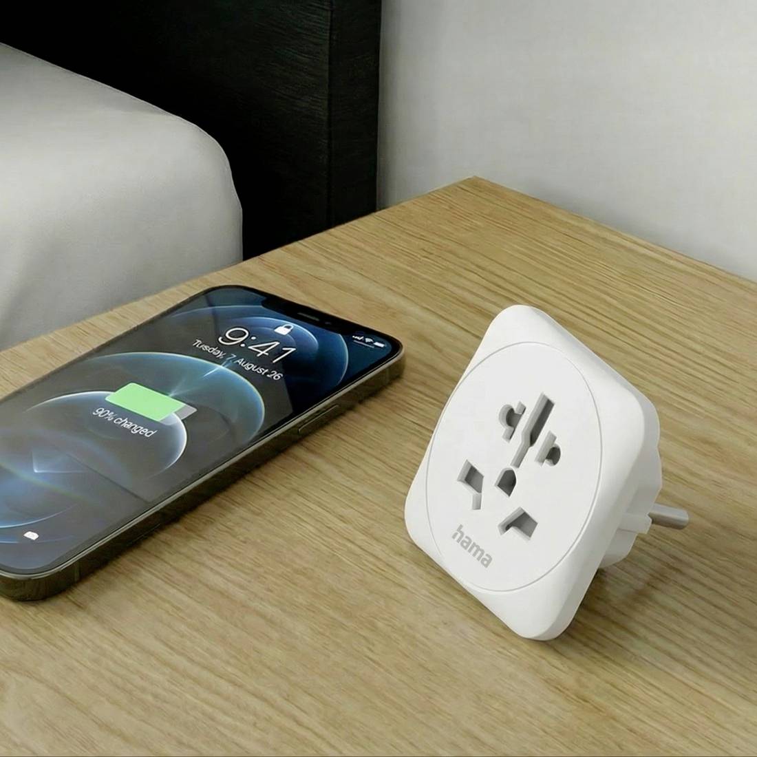Ein Smartphone lädt auf einem Holztisch neben einem weißen universellen Reiseadapter-Stecker auf.