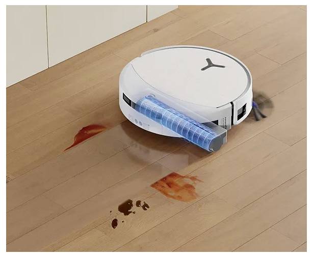 Roboterstaubsauger reinigt verschüttete Flüssigkeit auf einem Holzboden, wobei eine blaue Walzenbürste unter der vorderen Kante sichtbar ist.