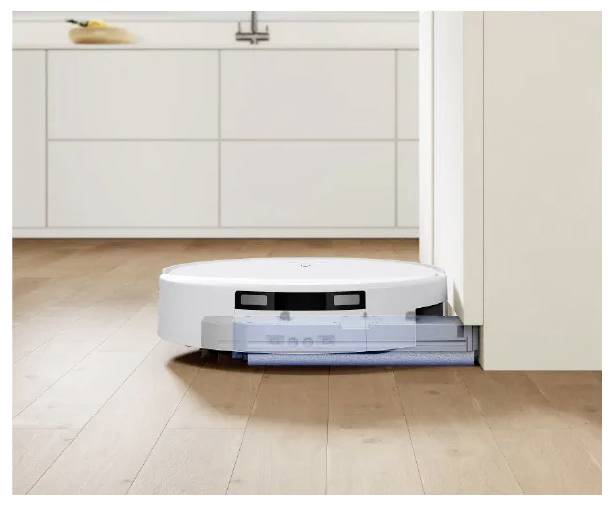 Roboterstaubsauger reinigt einen Holzküchenflur, während er sich neben einem Schrank in einer modernen weißen Küche andockt.