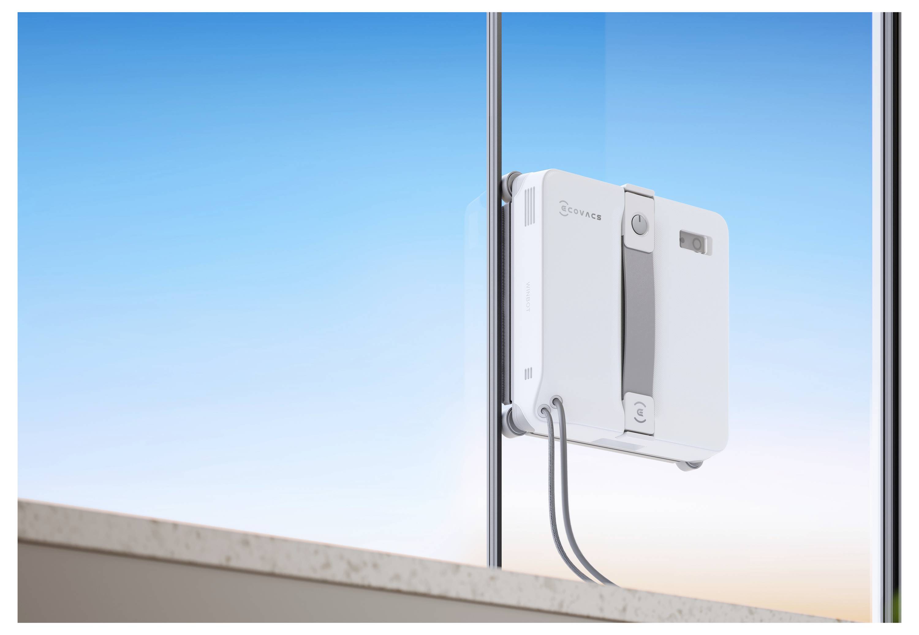 Weißes wandmontiertes Gerät, das an einer Glasplatte angebracht ist, wahrscheinlich ein Fensterreinigungs- oder Reinigungsroboter, mit sichtbaren Kabeln vor blauem Himmel.