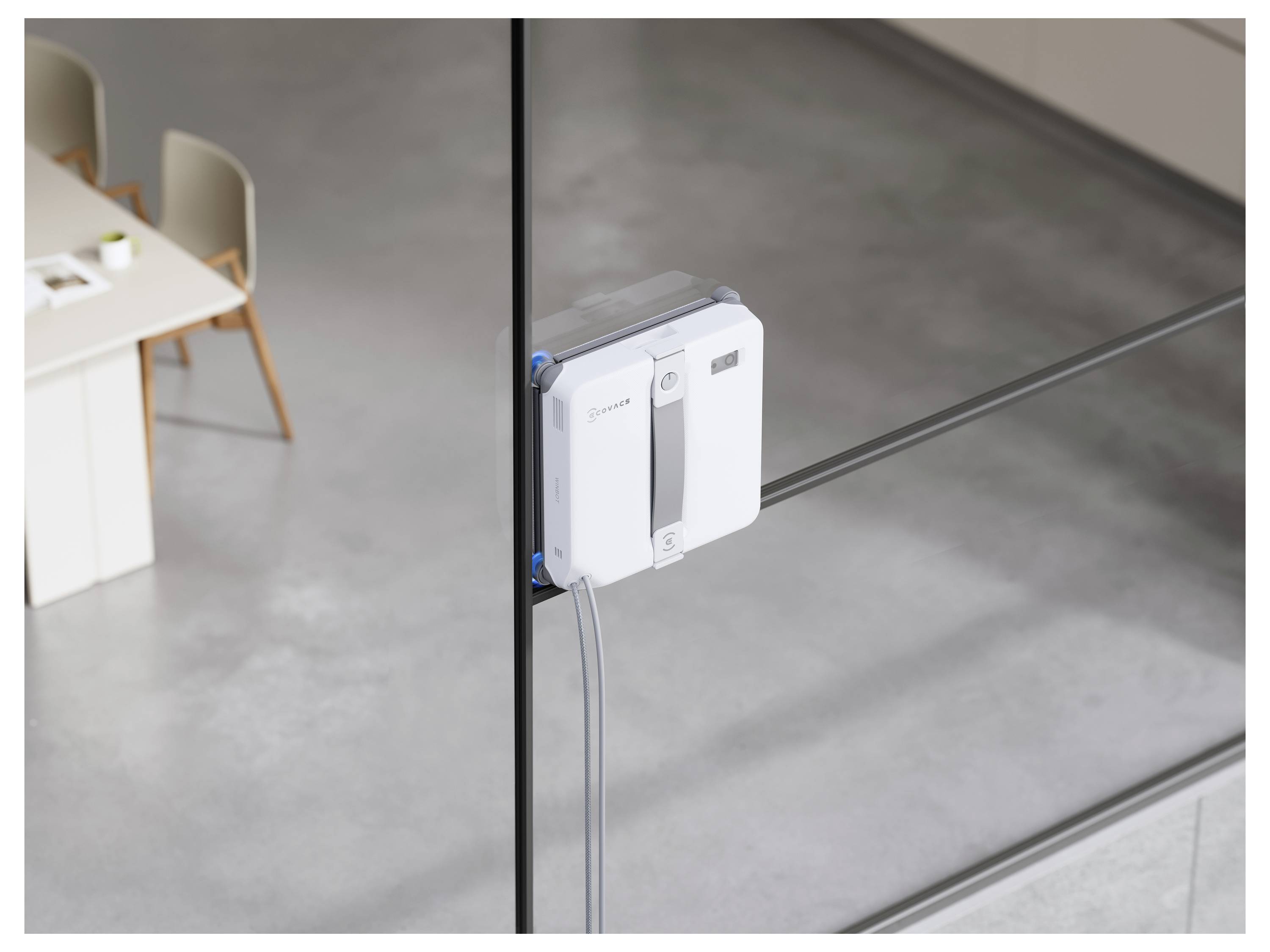 Fensterreinigungsroboter an einer innenliegenden Glaswand befestigt, mit Bürostühlen und einem Tisch im Hintergrund sichtbar.