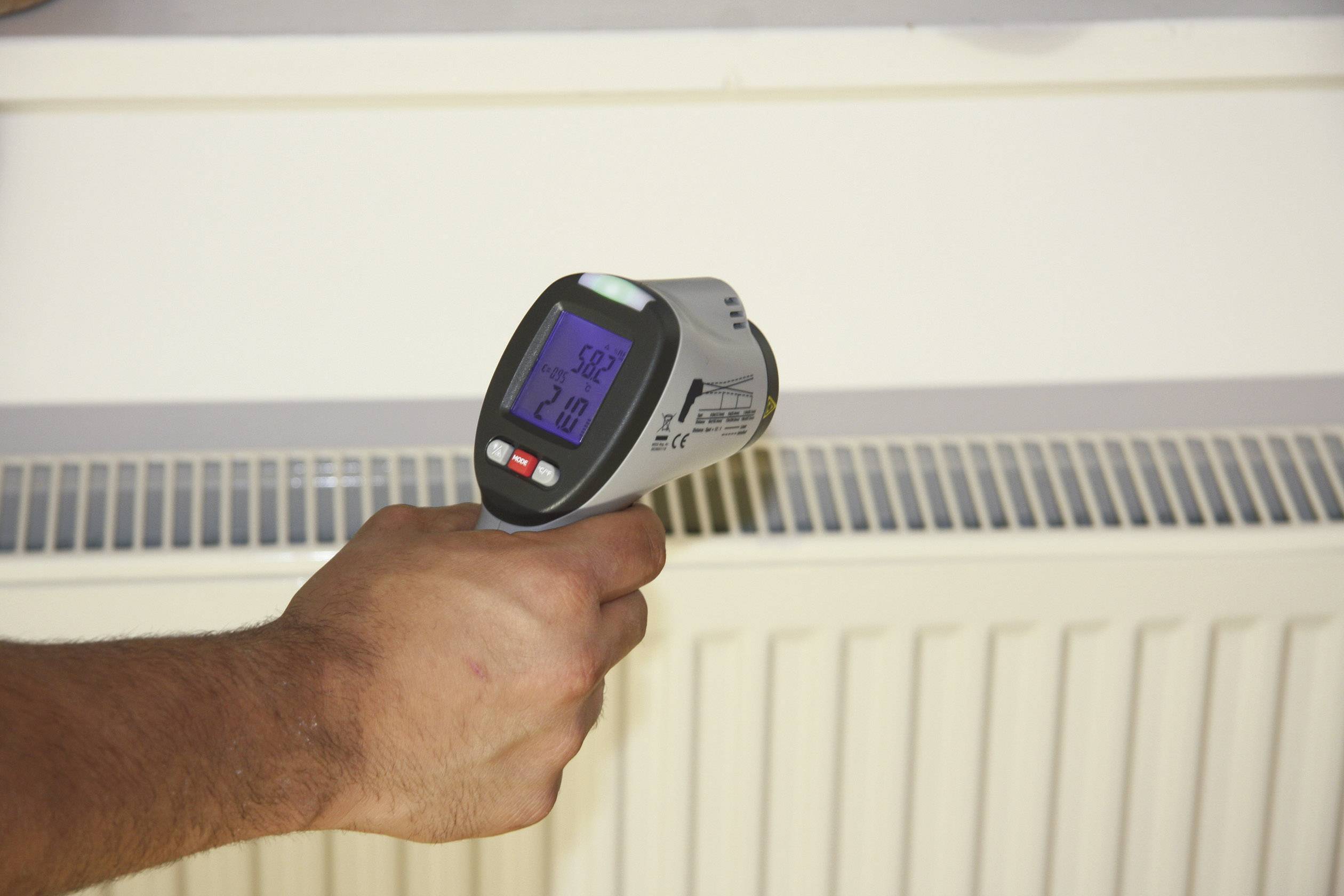 Eine Hand hält ein Infrarot-Thermometer, das die Temperatur eines Heizkörpers misst. Das Display zeigt 52,3 Grad Celsius.