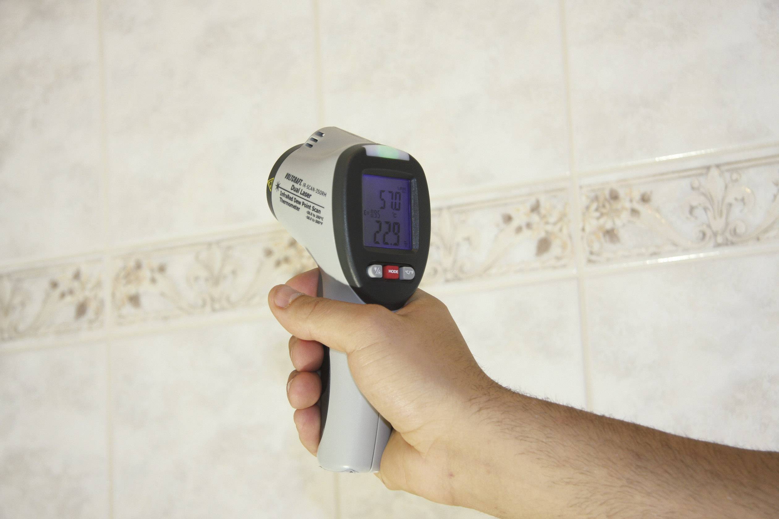 Eine Hand hält ein Infrarot-Thermometer vor eine geflieste Wand. Das Display zeigt Temperaturmessungen von 5,0 und 22,9 Grad.