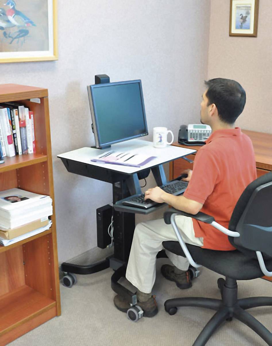 Ein Mann sitzt an einem Schreibtisch und arbeitet am Computer. Neben ihm steht ein Bücherregal. Büromaterialien auf dem Tisch.