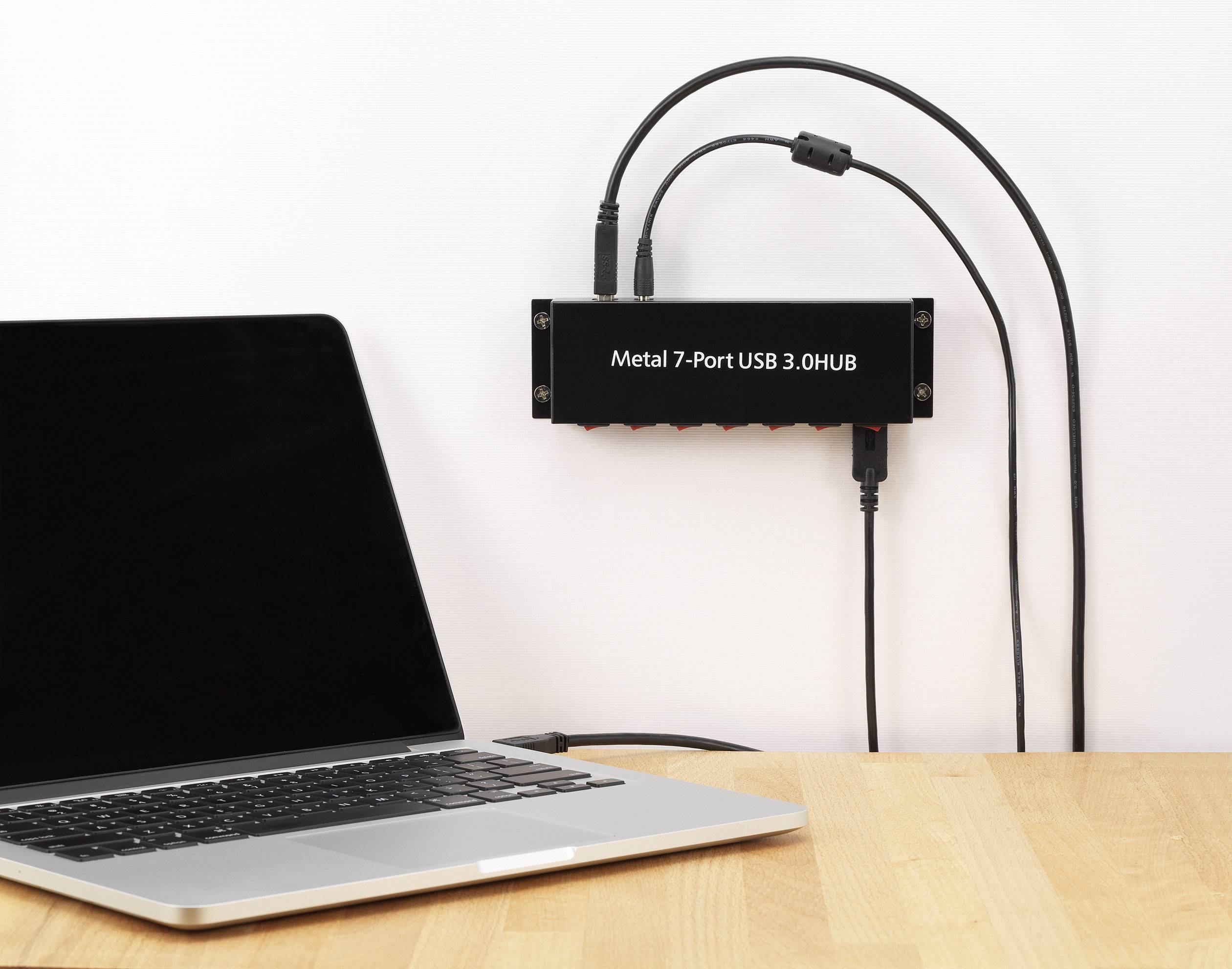 Laptop auf Holztisch verbunden mit einem schwarzen Metall-USB-Hub mit sieben Ports. Zwei USB-Kabel sind angeschlossen.