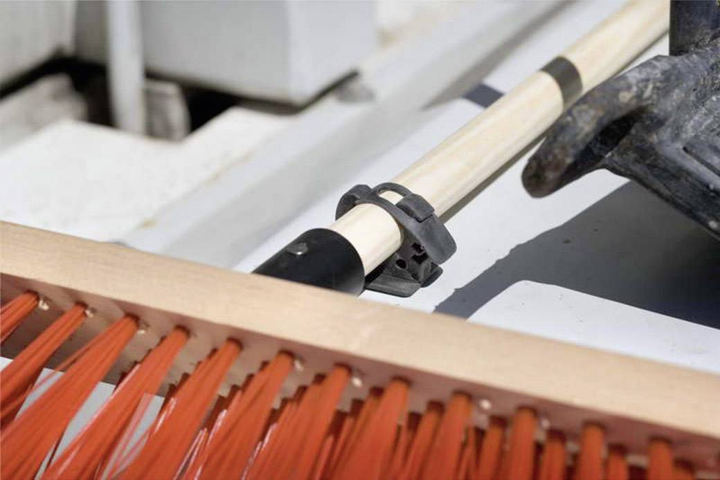 Ein Besen mit orangefarbenen Borsten liegt auf einem Holzgriff, der mit einer Halterung befestigt ist, vor einem unscharfen Hintergrund.