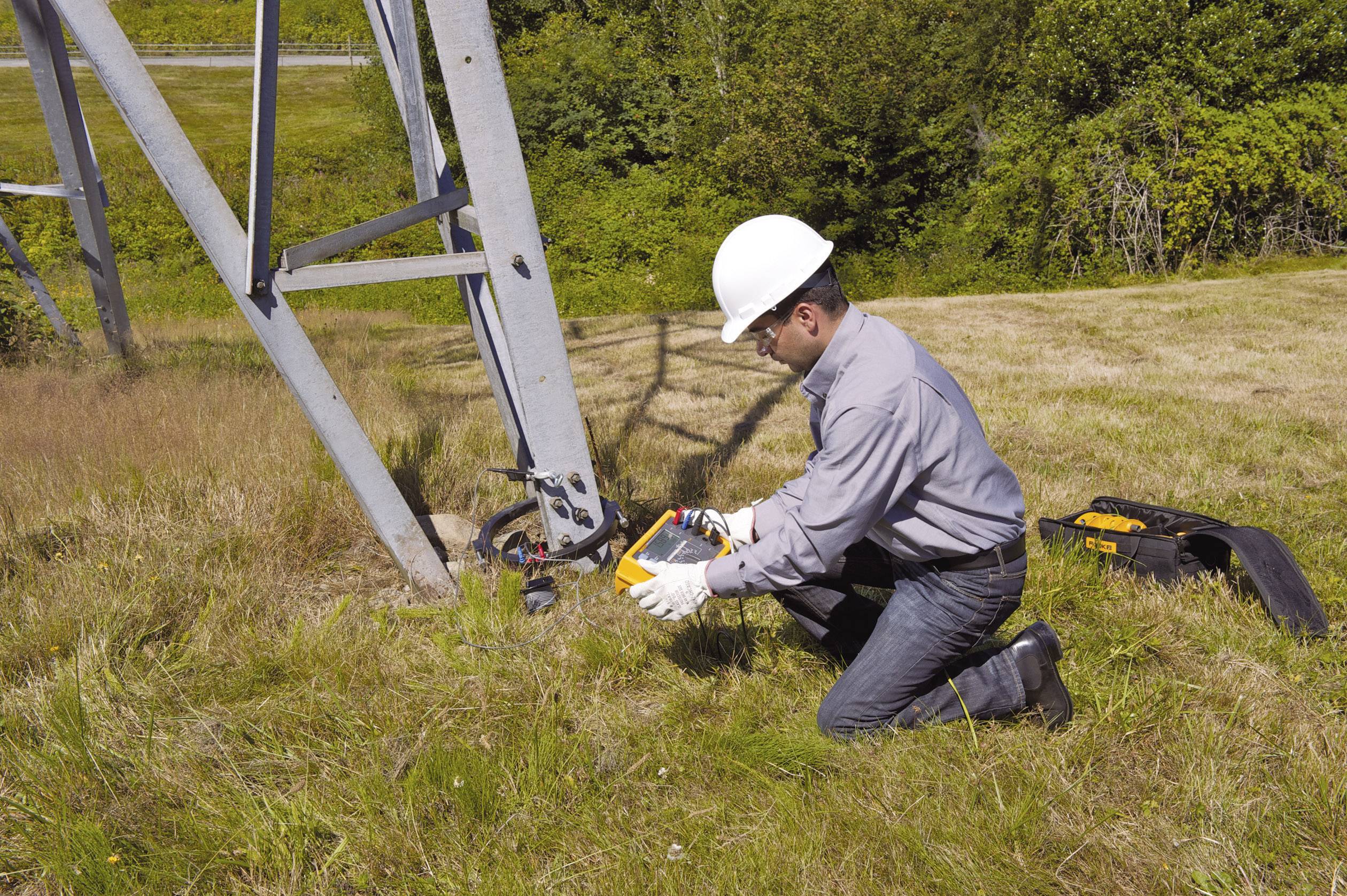 Ein Arbeiter mit Helm kniet auf Gras und nutzt ein Messgerät an einem Metallmast zur Wartung oder Überprüfung technischer Strukturen.