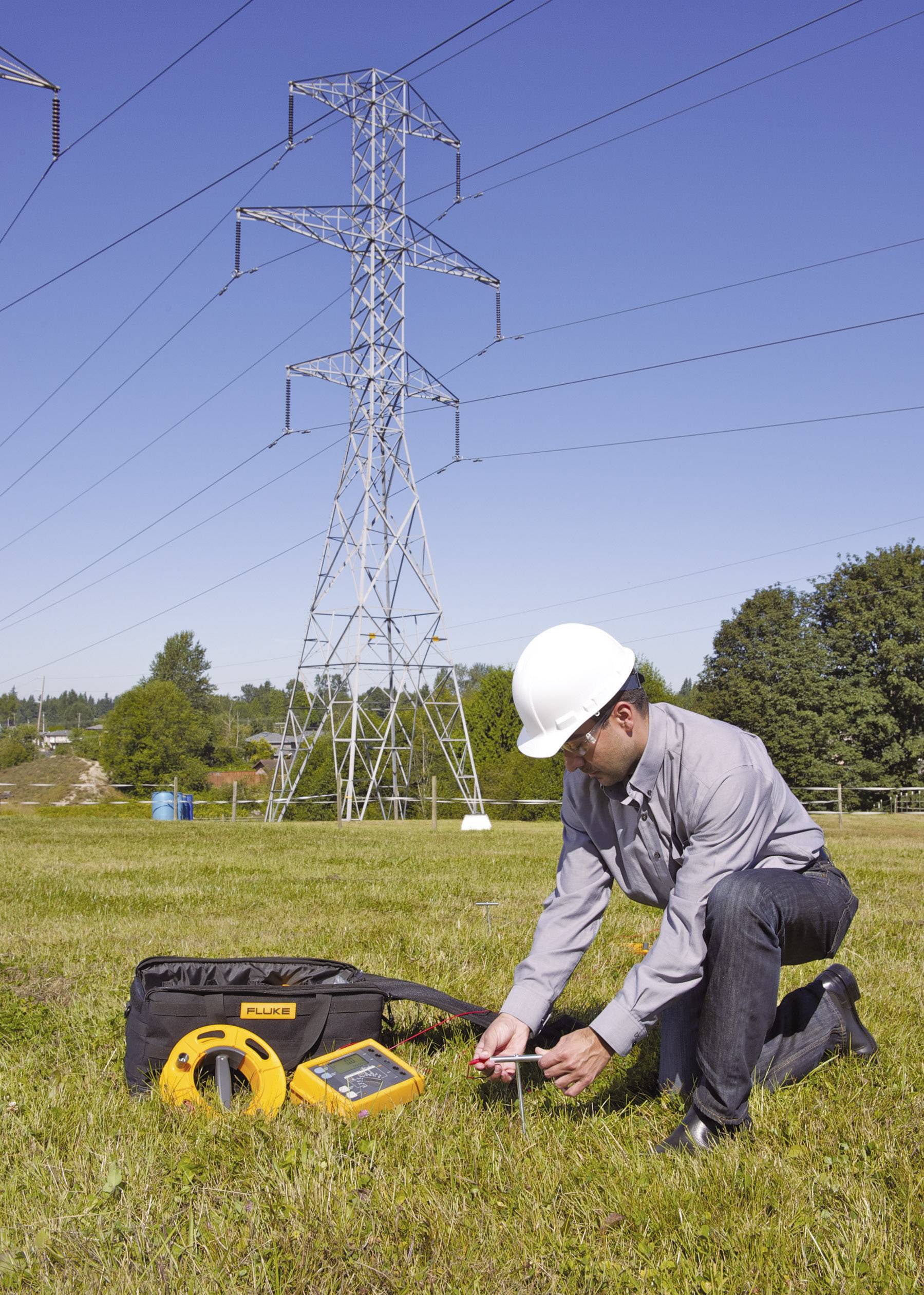 Ein Arbeiter in Schutzhelm misst elektrische Spannung mit einem Messgerät auf einem Feld. Im Hintergrund ein Strommast.