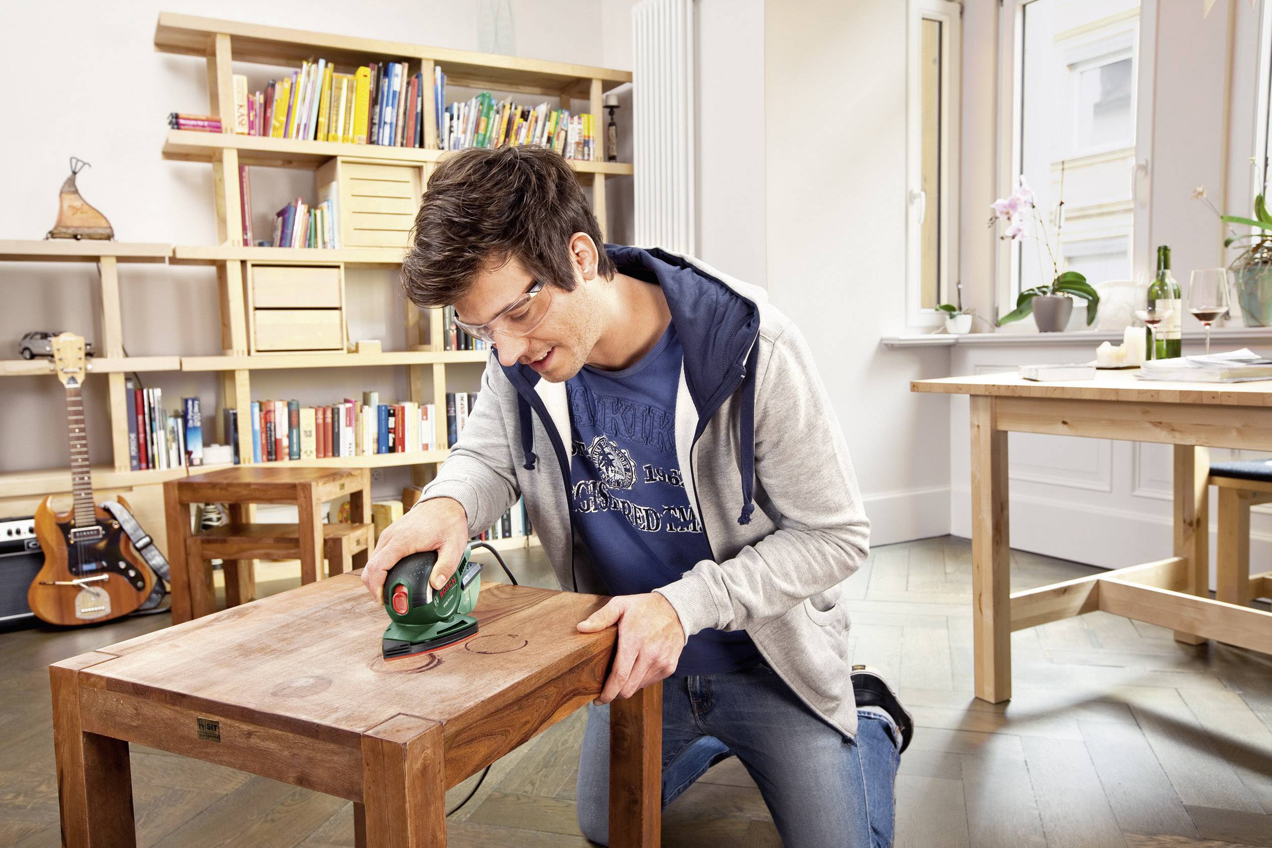 Ein Mann schleift mit einem Werkzeug das Holz eines kleinen Tisches in einem hellen Zimmer mit Regalen und Tisch im Hintergrund.