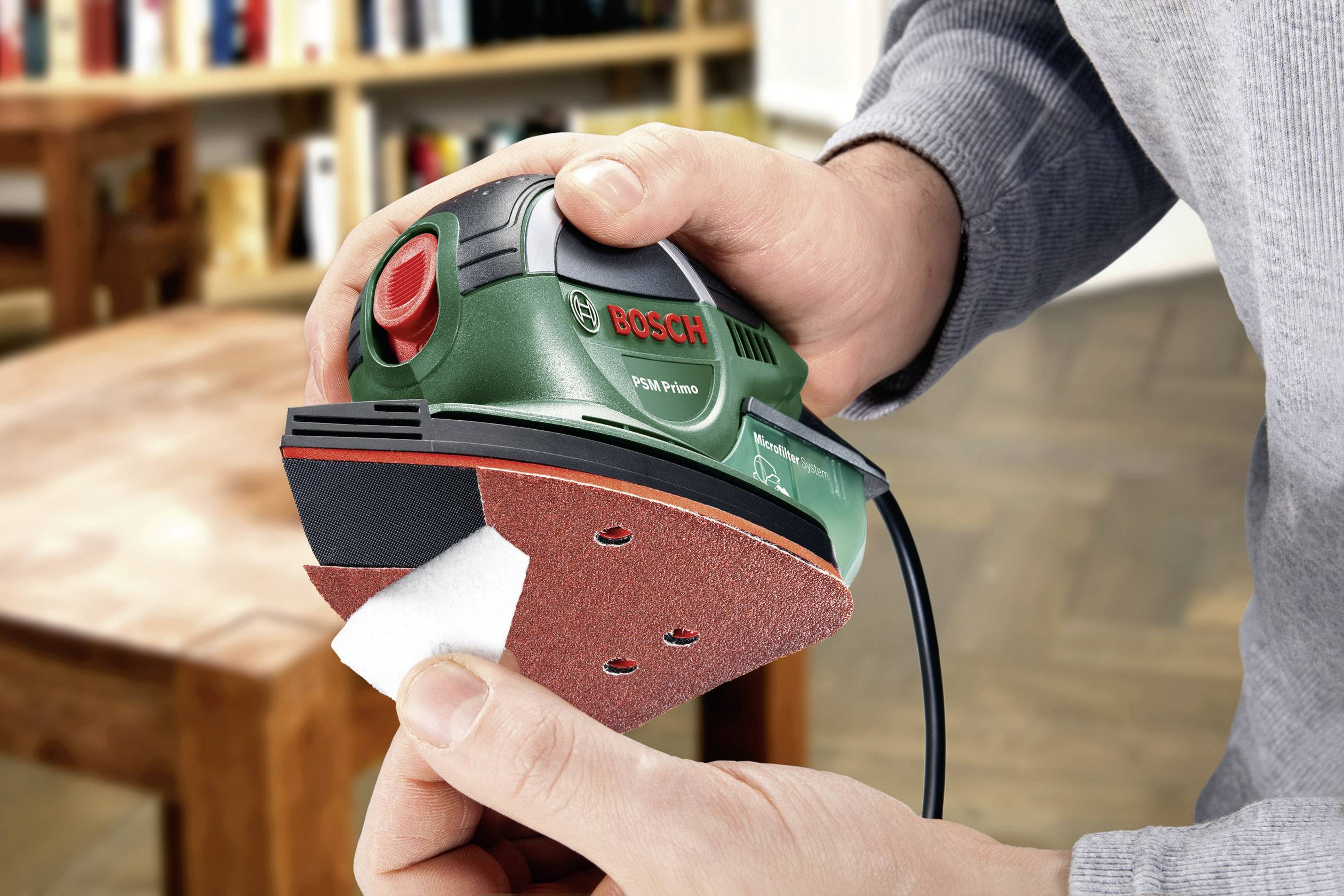 Eine Person hält einen grünen Bosch-Multischleifer und wechselt das Schleifpapier. Im Hintergrund ist ein Holztisch sichtbar.