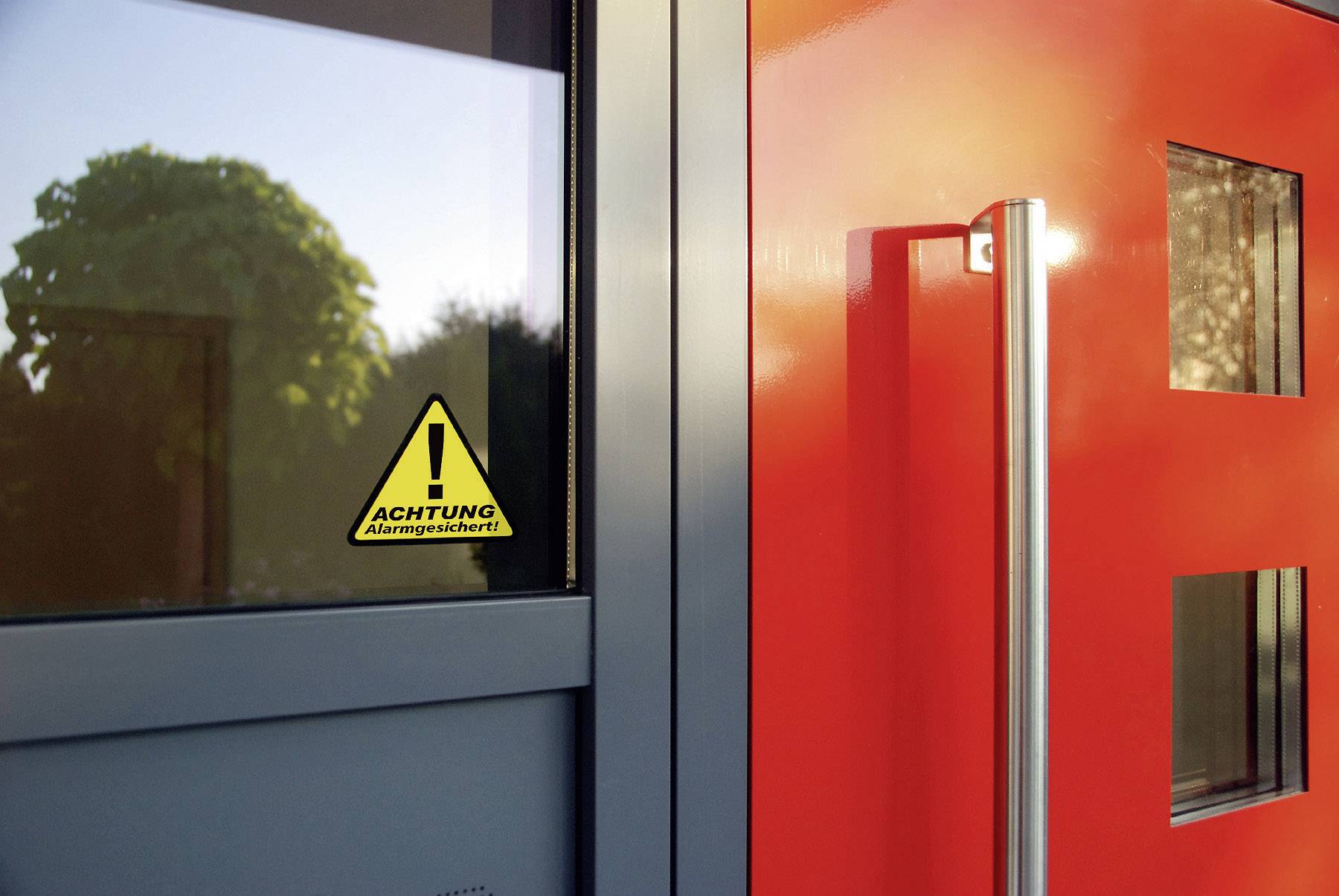 Eine rote Tür mit Glasfenstern und einer Fensterscheibe daneben. Auf der Scheibe ist ein gelbes Warnschild mit 'Achtung Alarmgesichert'.