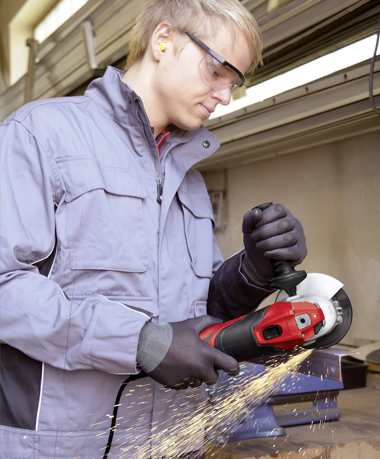 Ein Mann mit Schutzbrille und Handschuhen schleift Metall mit einem Winkelschleifer in einer Werkstatt. Funken fliegen vom Arbeitsstück.