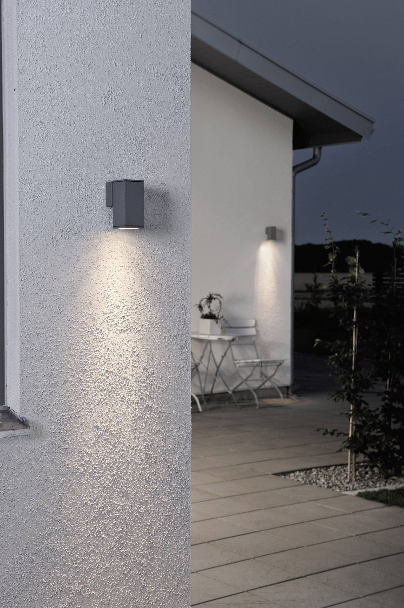 Außenansicht eines Hauses bei Dämmerung mit zwei Wandlampen und einem Tisch mit Stühlen im Hintergrund. Beleuchteter Weg führt dorthin.