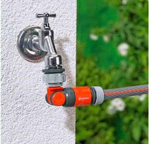 Ein Wasserhahn an einer weißen Wand mit einem angeschlossenen Gartenschlauch, der nach rechts führt. Im Hintergrund ist grüne Vegetation.