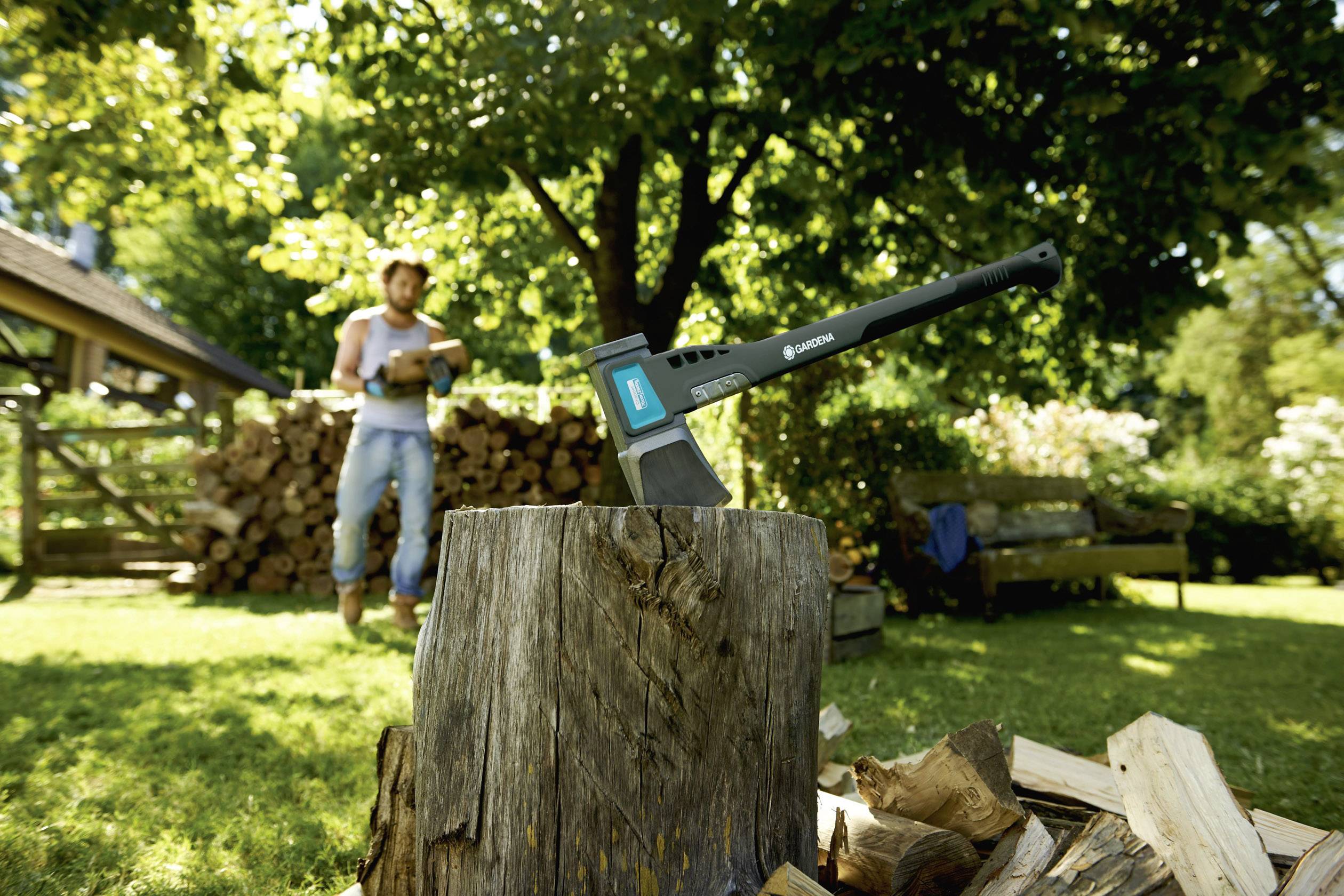 Eine Axt steckt in einem Baumstumpf in einem sonnigen Garten. Im Hintergrund steht eine Person vor einem Stapel Holz.