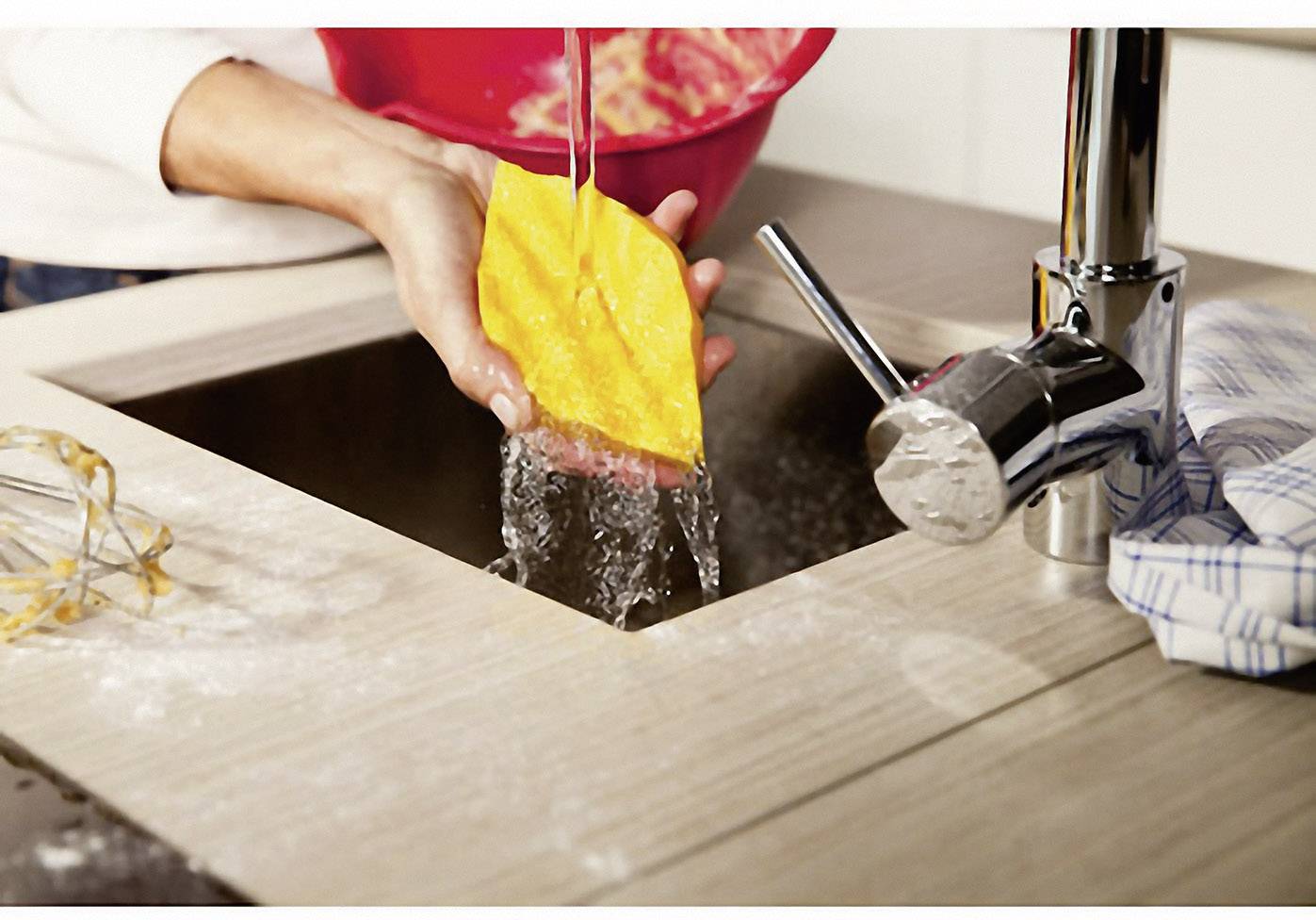 Person wäscht einen gelben Schwamm unter fließendem Wasser in der Küche, umgeben von einem Schneebesen und einem gestreiften Tuch.