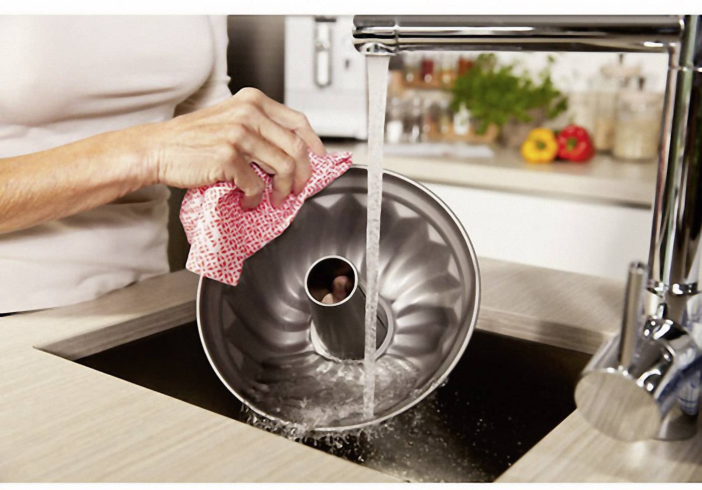 Person spült Kuchenbackform unter fließendem Wasser im Waschbecken ab. Im Hintergrund sind bunte Paprika in der Küche zu sehen.