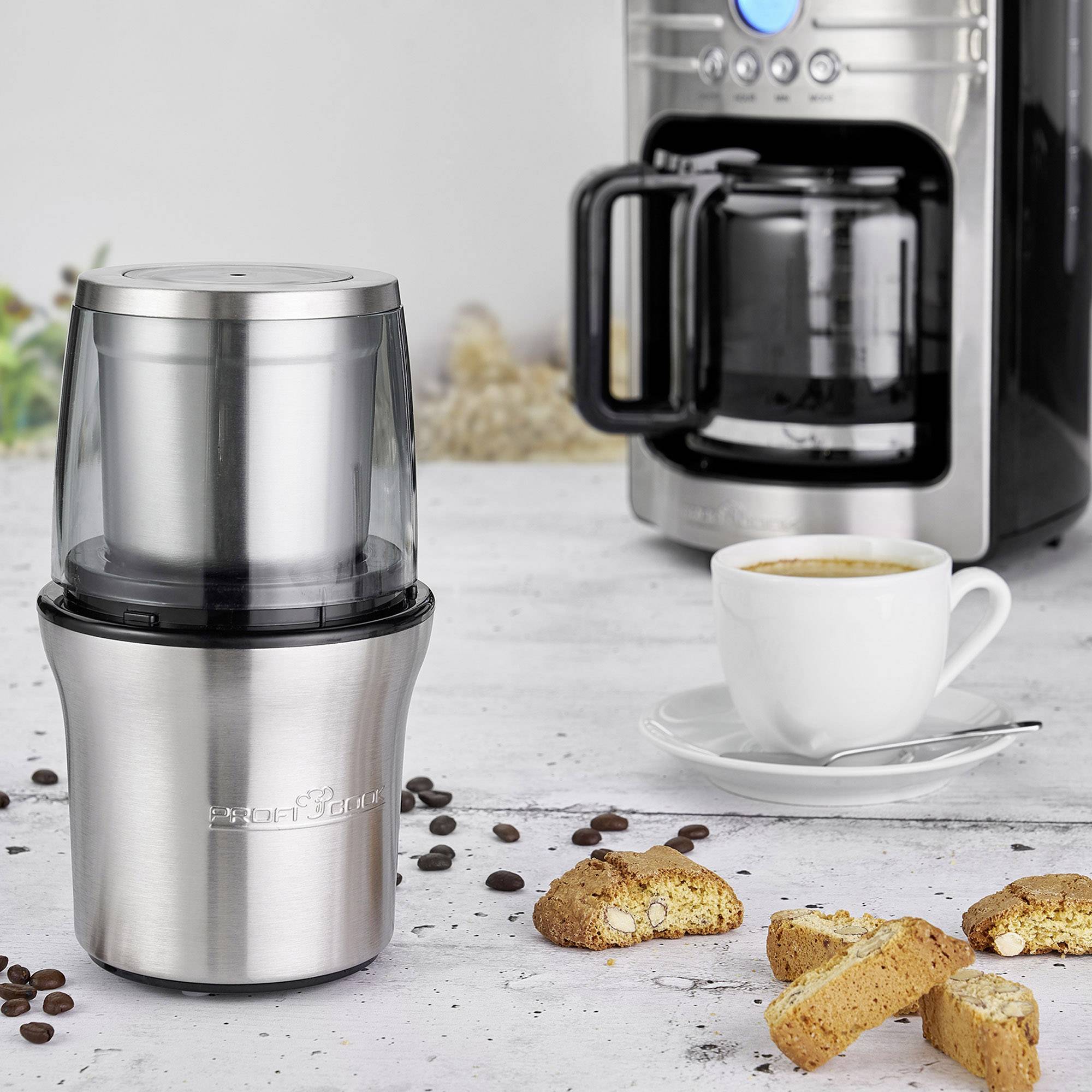 Ein elektrischer Kaffeemühlenbehälter steht auf einem Tisch neben einer Tasse Kaffee, einer Kaffeemaschine und Keksen.