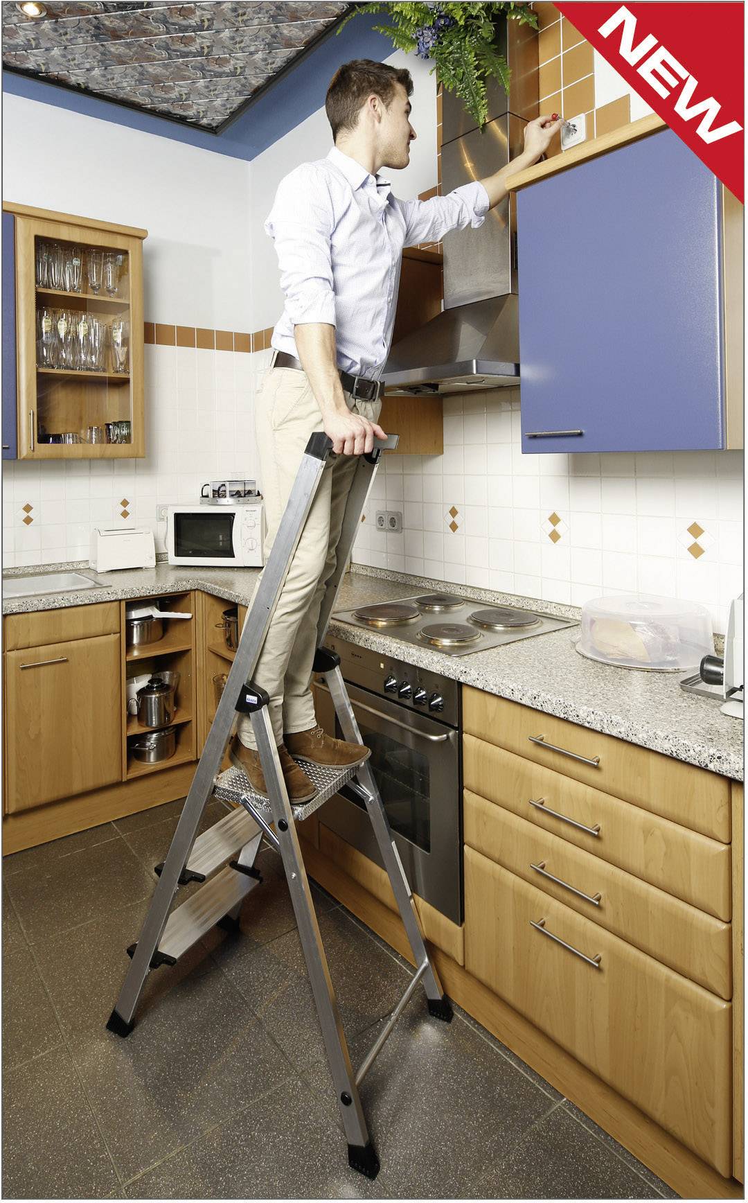 Ein Mann steht auf einer Klappleiter in einer Küche und überprüft eine Rauchmelder an der Decke. Links im Bild befinden sich Küchenschränke.