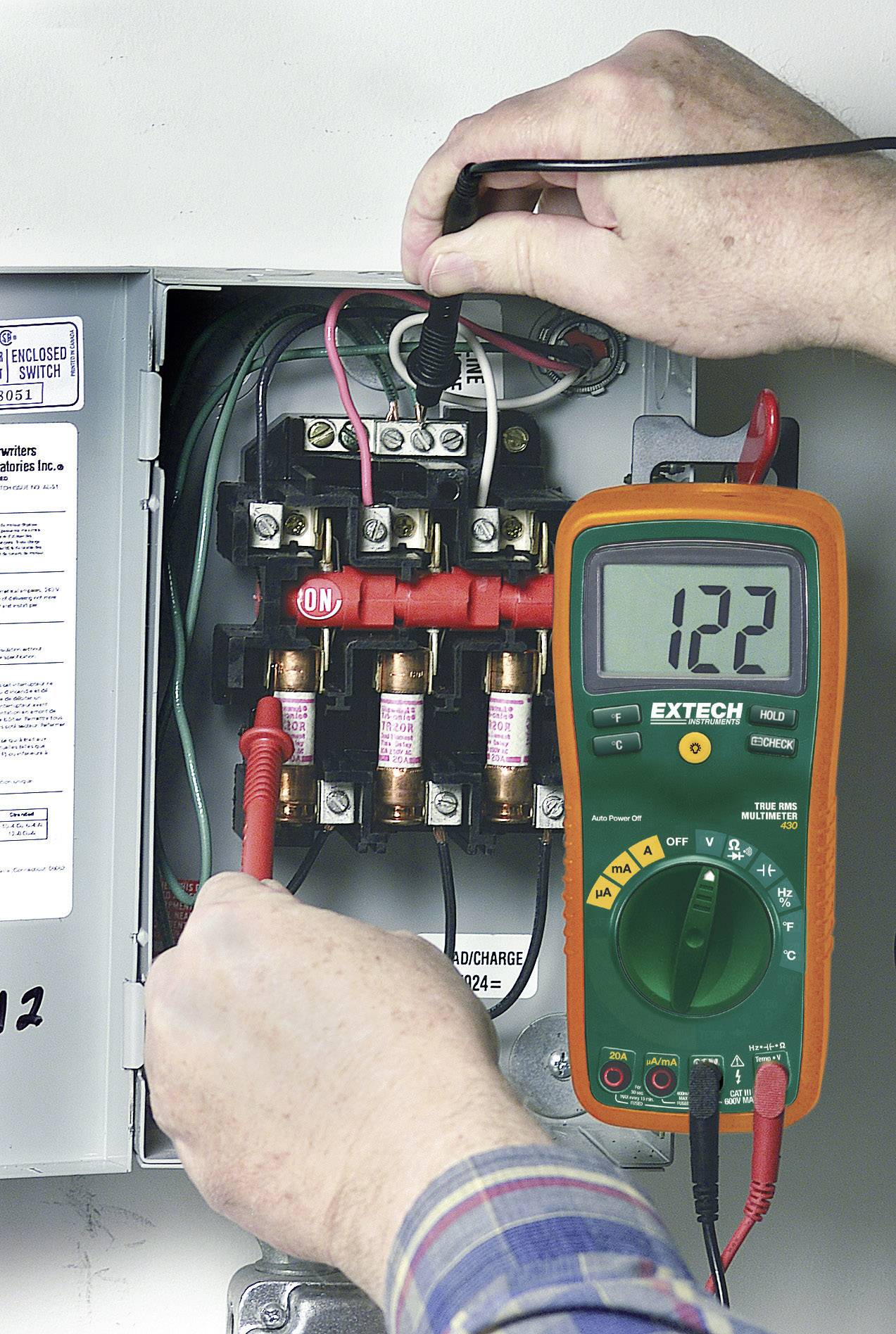 Ein Elektriker misst die Spannung mit einem Multimeter an einem geöffneten Sicherungskasten. Das Display zeigt '122'.