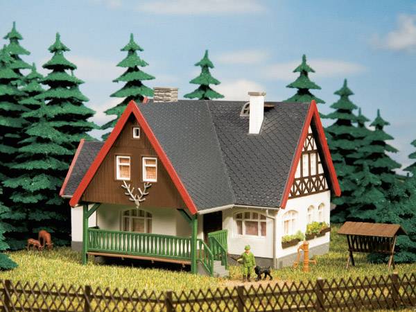 Ein kleines Modellhaus mit Giebeldach, Veranda und Fachwerkdetails steht umgeben von Tannenbäumen und einem Holzzaun in ländlicher Umgebung.