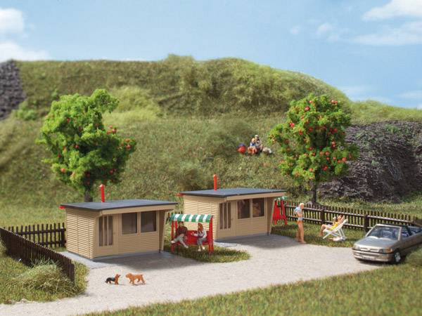 Zwei kleine Holzhäuser in einem grünen Ferienpark, umgeben von Bäumen mit roten Äpfeln. Ein Auto parkt neben den Häusern.