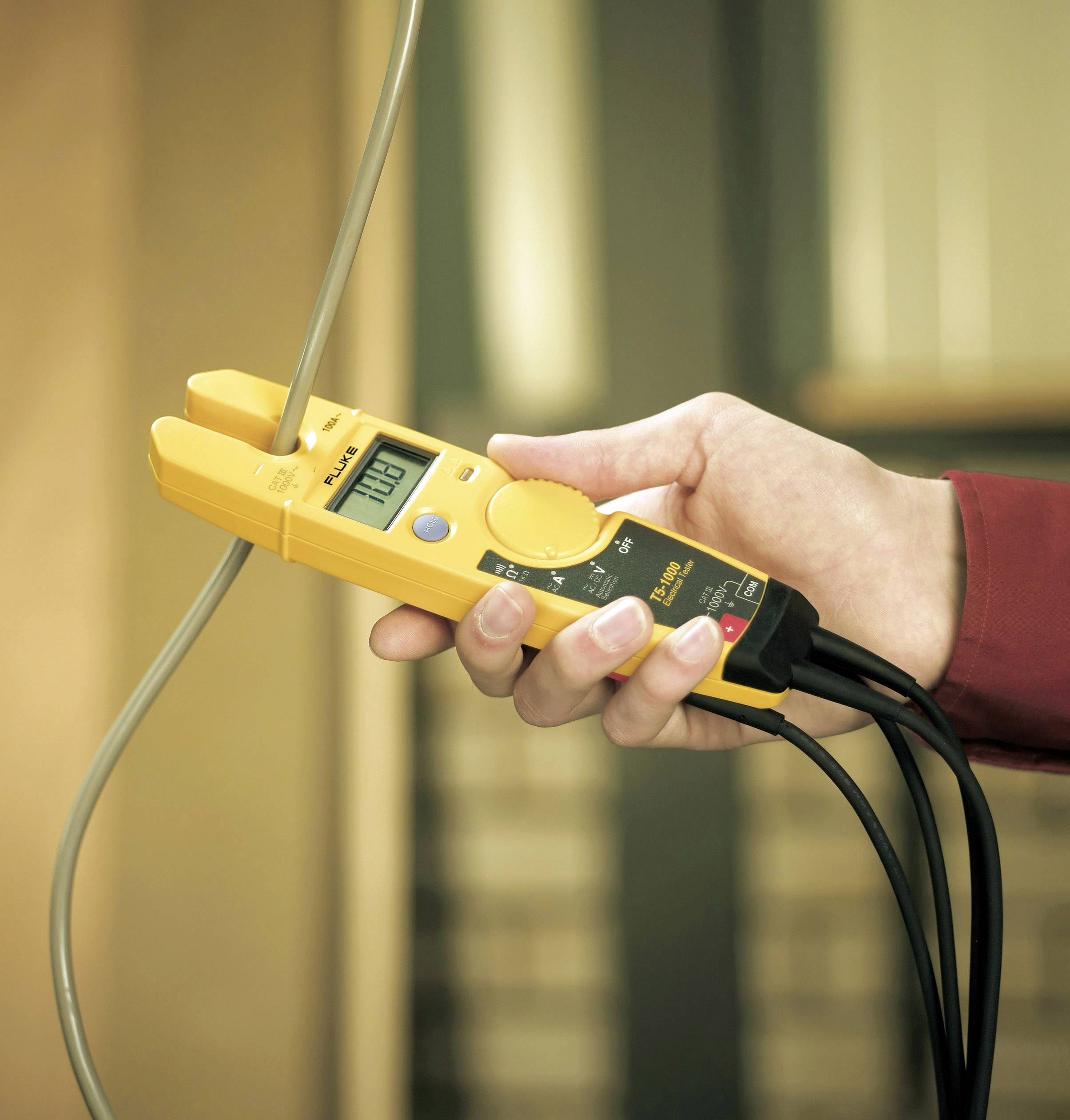 Eine Hand hält ein gelbes elektrisches Messgerät, das eine Spannung von 120,2 Volt anzeigt und mit Kabeln verbunden ist.