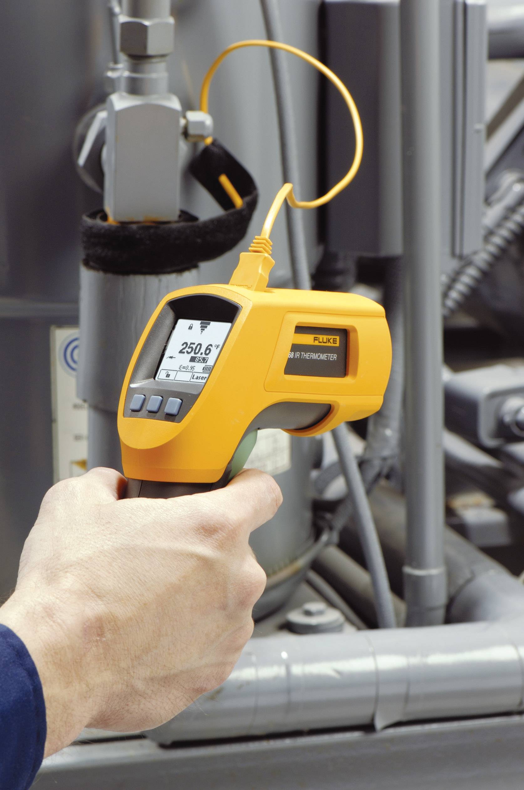 Eine Hand hält ein Infrarot-Thermometer, das eine Temperatur von 25,6 Grad Celsius misst, vor einer industriellen Maschine.