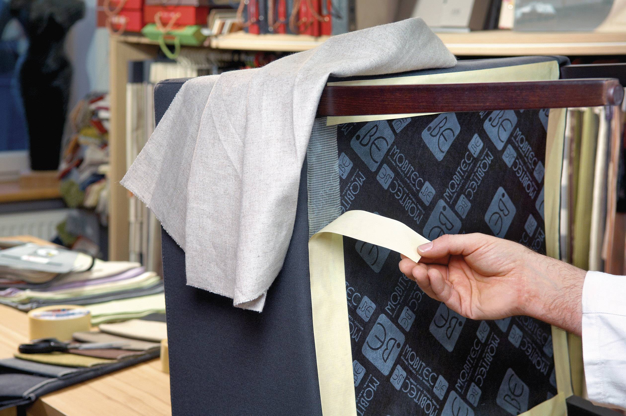 Eine Hand zeigt Stoffmuster auf einer gepolsterten Oberfläche, umgeben von verschiedenen Stoffen und Materialien in einem Raum.