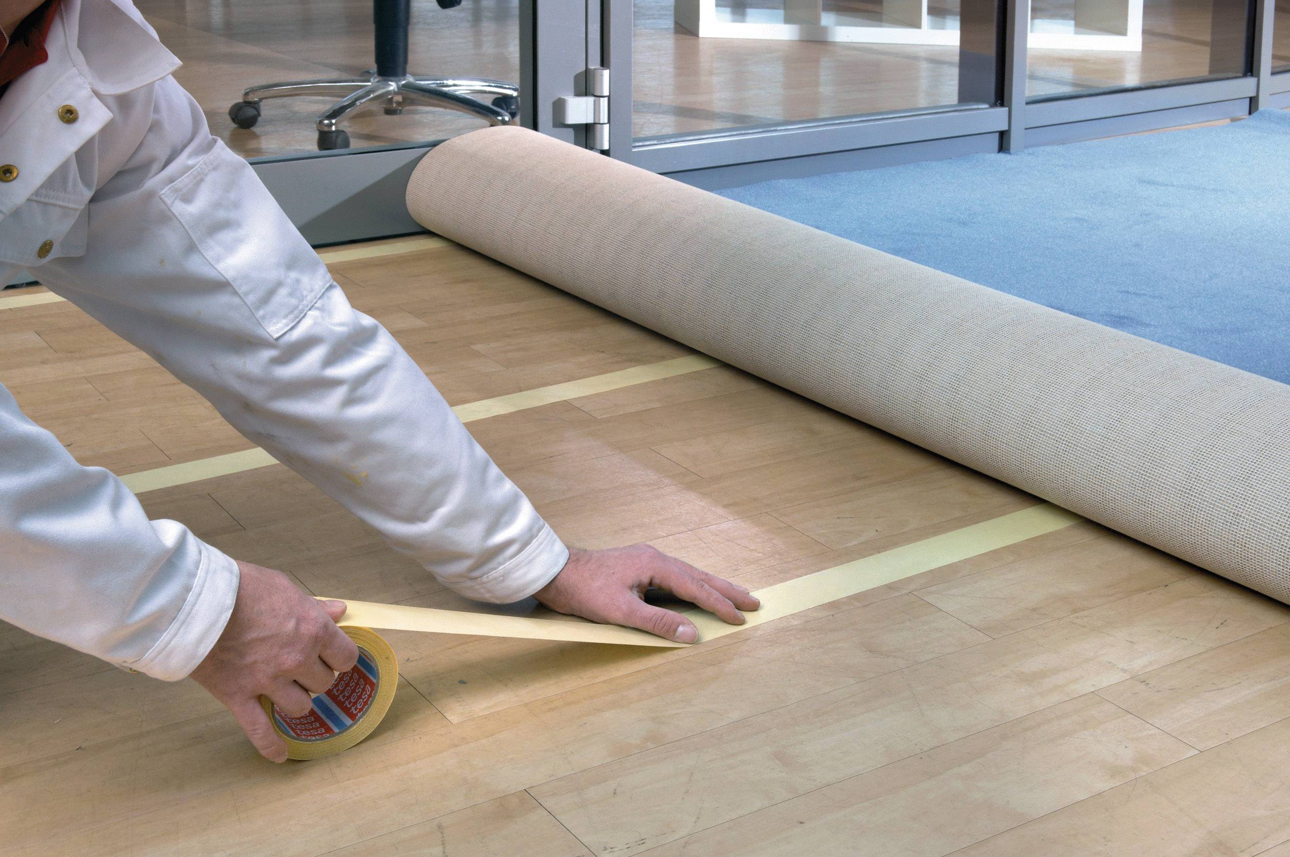 Eine Person klebt Klebeband auf einen Holzboden, um einen Teppich oder Läufer auszurollen, in einem Büro mit Glaswänden.