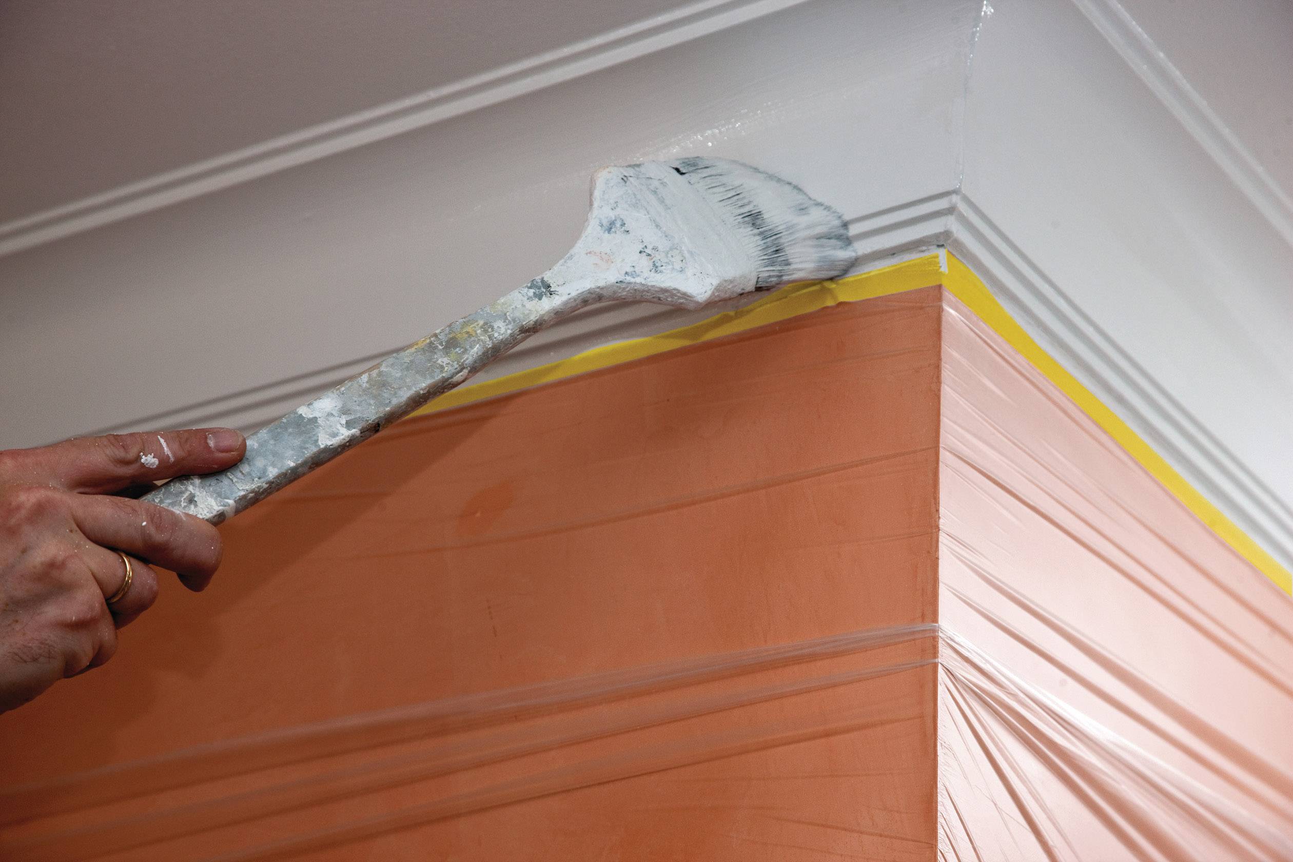 Ein Pinsel trägt weiße Farbe auf die oberste Kante einer orange gestrichenen Wand auf. Eine gelbe Linie dient als Markierung.