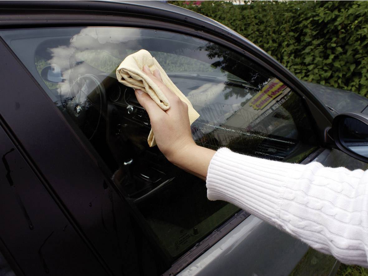 Eine Person reinigt das Autofenster von außen mit einem beigen Tuch. Das Auto steht im Freien, bewölkter Himmel spiegelt sich im Glas.