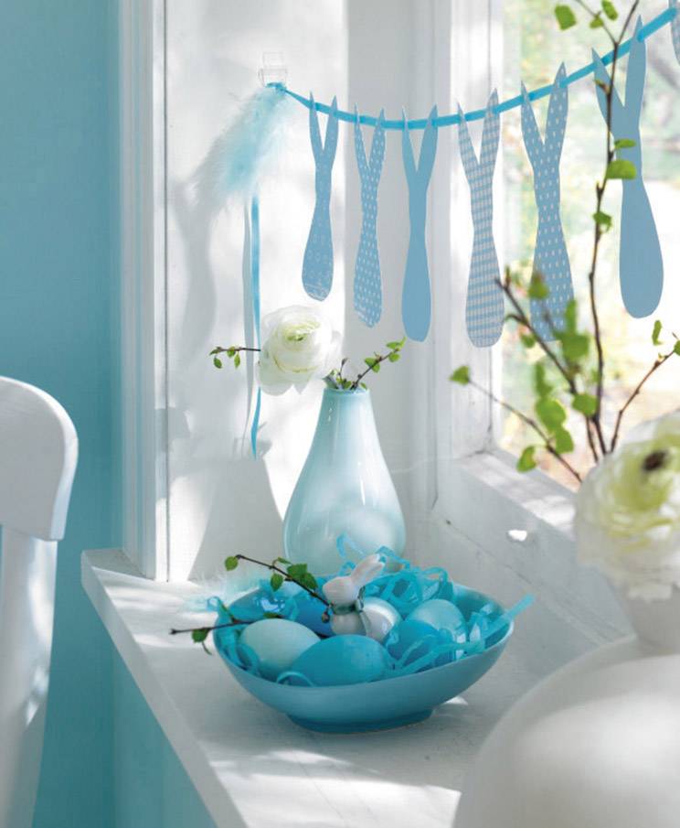 Ein Fensterbrett mit blauen Dekorationen: eine Vase mit weißen Blumen, eine Schüssel mit blau gefärbten Eiern und ein blauer Wimpel.