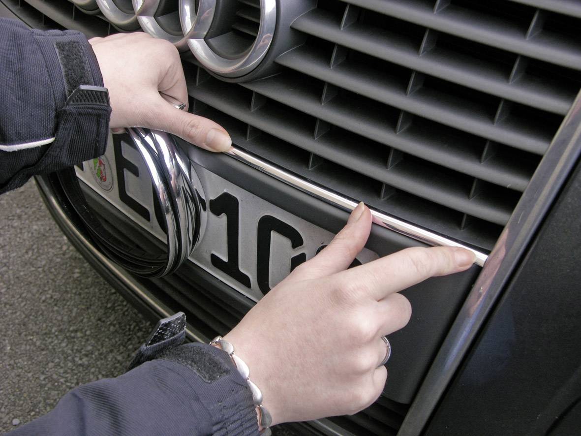 Eine Person befestigt ein ovales, verchromtes Objekt an der Vorderseite eines Autos nahe einem Nummernschild.