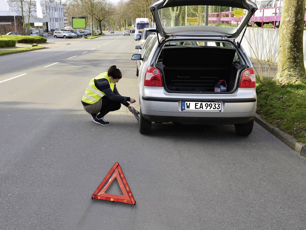 Eine Frau wechselt bei offenem Kofferraum auf einer Straße einen Reifen an einem Auto. Ein Warndreieck ist aufgestellt.