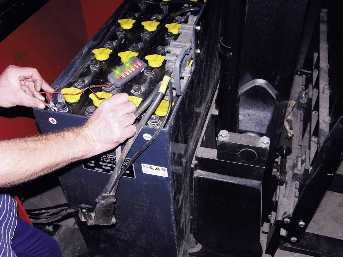 Eine Person prüft eine Batterie in einem industriellen Gerät, umgeben von schwarzen und gelben Komponenten, mit einem Multimeter.