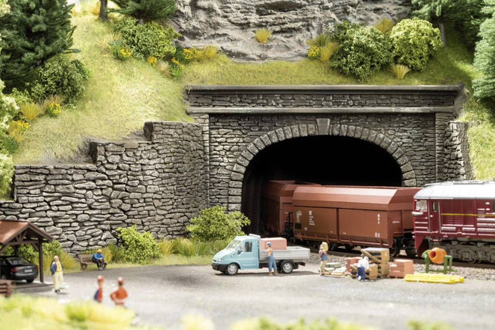 Ein Modellzug fährt aus einem steinernen Tunnel in einer Landschaft mit Bäumen und Figuren, die nahe einem kleinen, blauen Lastwagen stehen.