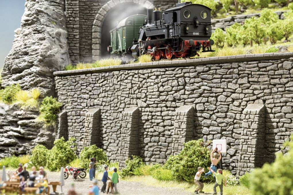 Eine schwarze Dampflok fährt aus einem Tunnel auf einer Modellbahnlandschaft, darunter spielen Figuren Basketball.