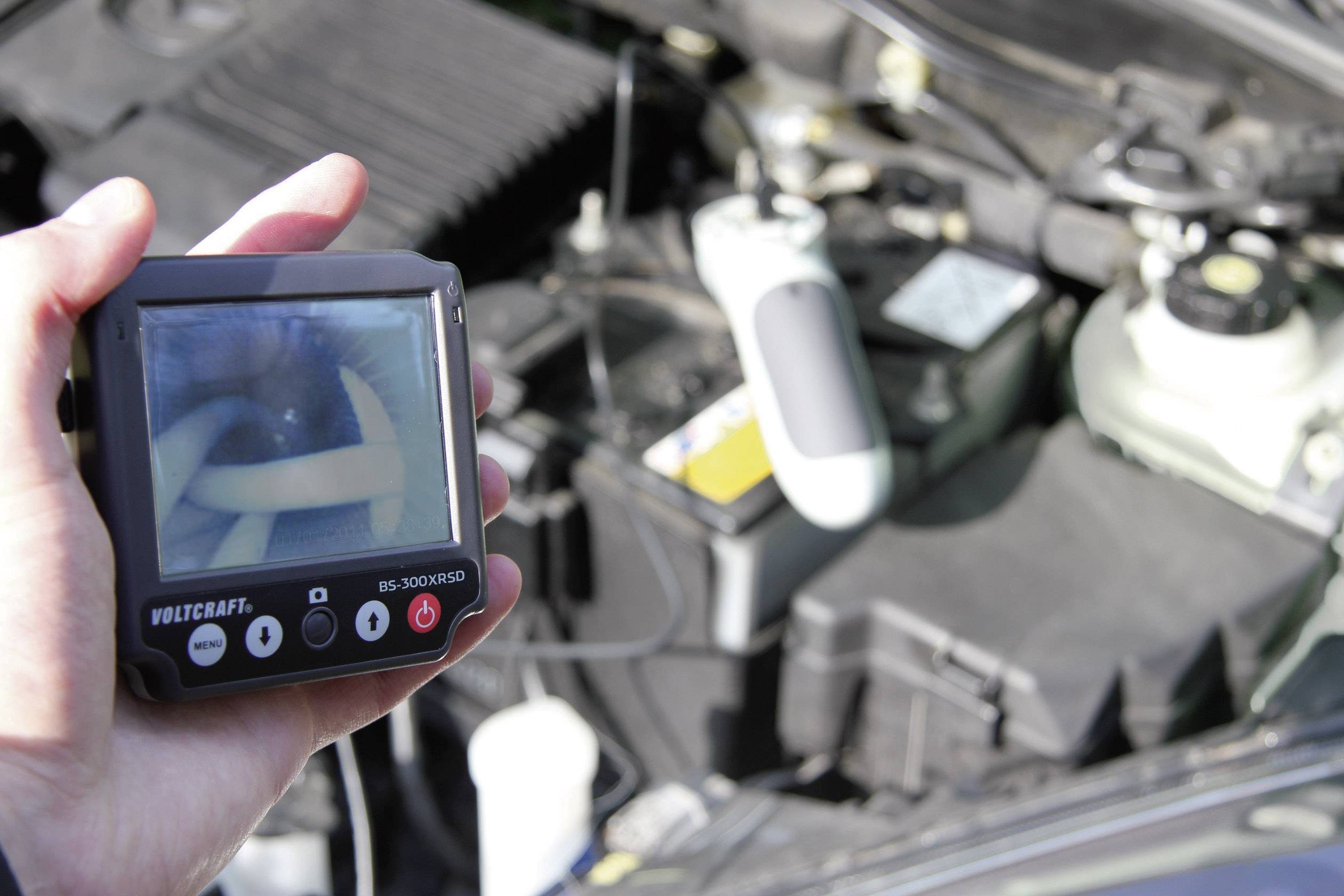 Eine Person hält ein Diagnosegerät mit Bildschirm, das den inneren Zustand eines Autoteils anzeigt. Im Hintergrund ist der Motorraum sichtbar.
