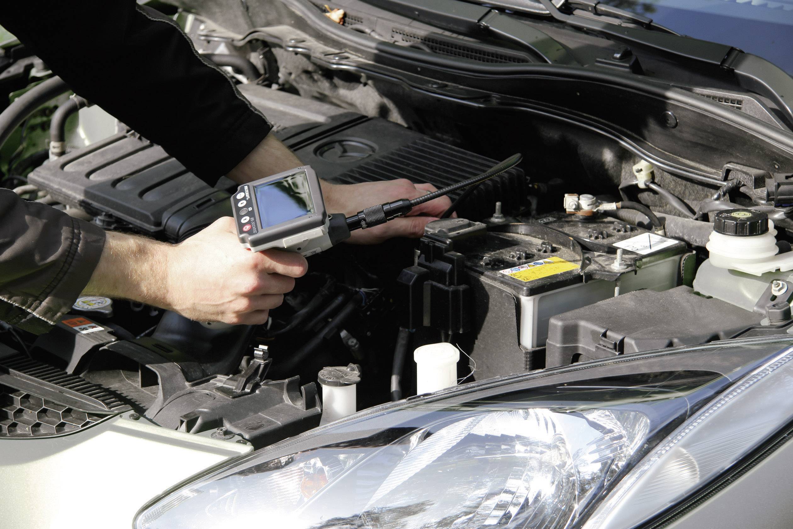 Eine Person überprüft den Motor eines Autos mit einem Diagnosegerät. Die Motorhaube ist geöffnet, und das Gerät zeigt Daten auf einem Bildschirm an.