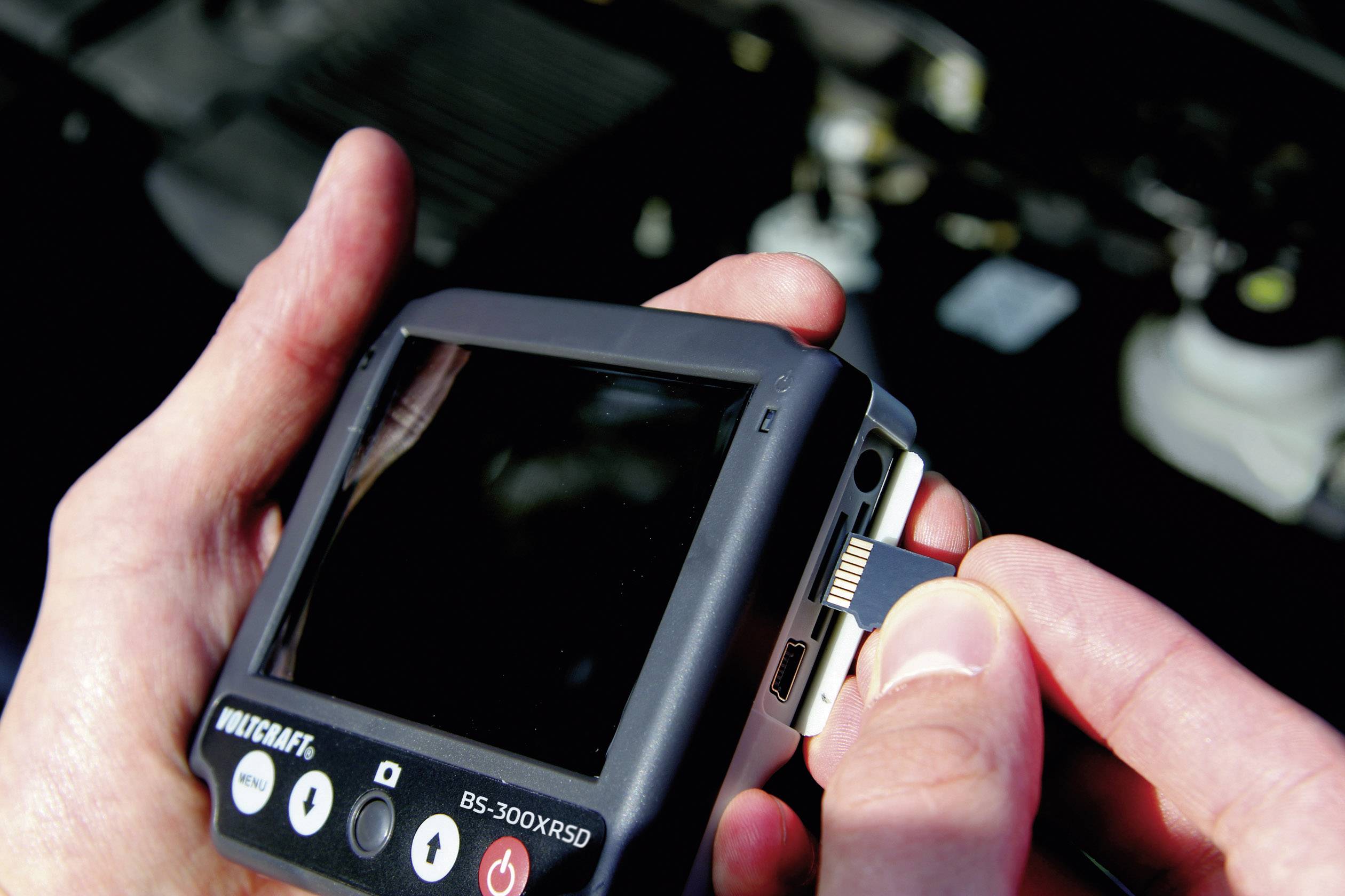 Eine Person steckt eine Speicherkarte in ein Diagnosegerät. Im Hintergrund sind verschwommene Autoteile zu sehen.