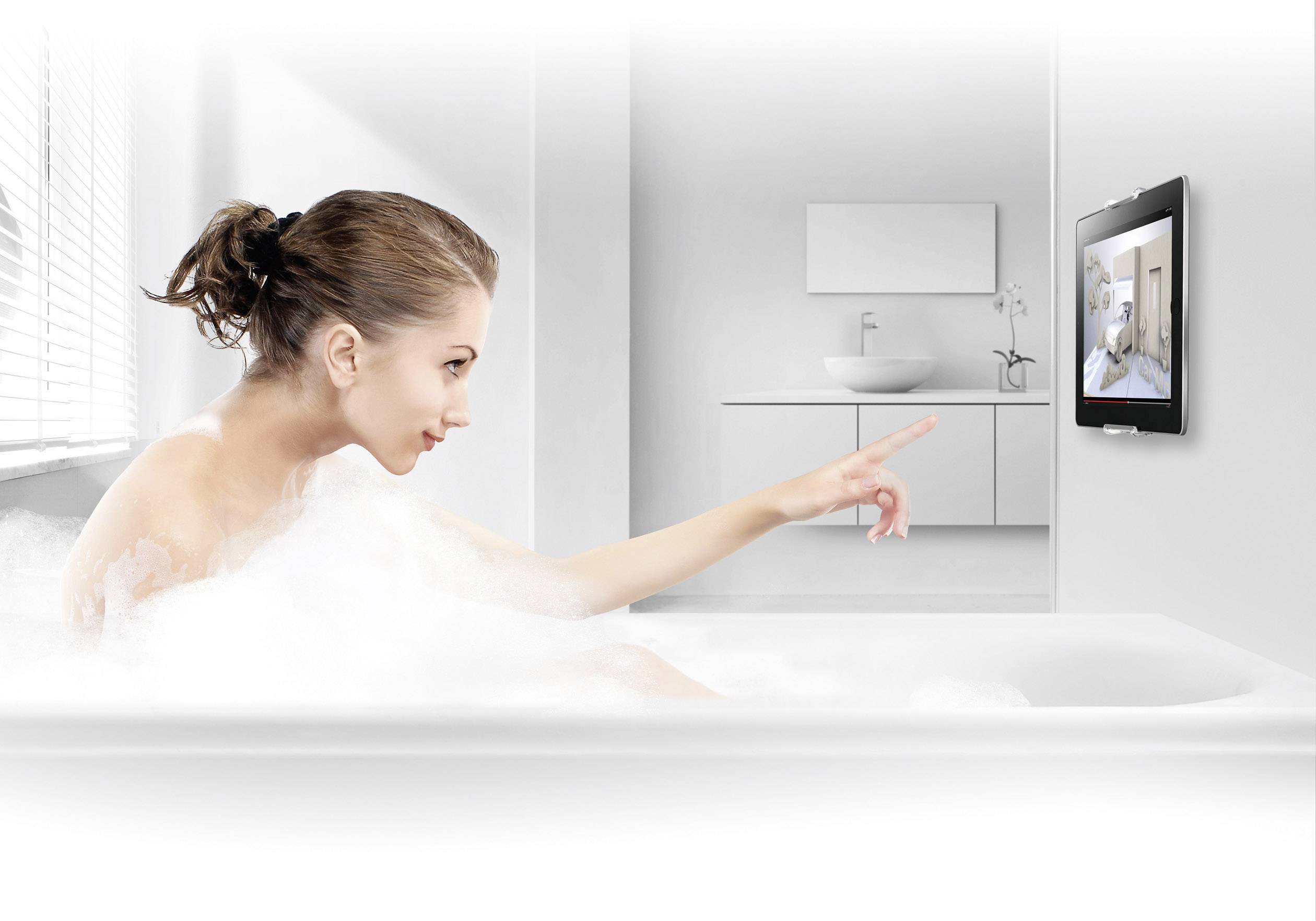 Eine Frau in einer Badewanne zeigt auf ein Tablet an der Wand, das ein Badezimmer anzeigt. Sie scheint interaktiv mit der Technik zu interagieren.