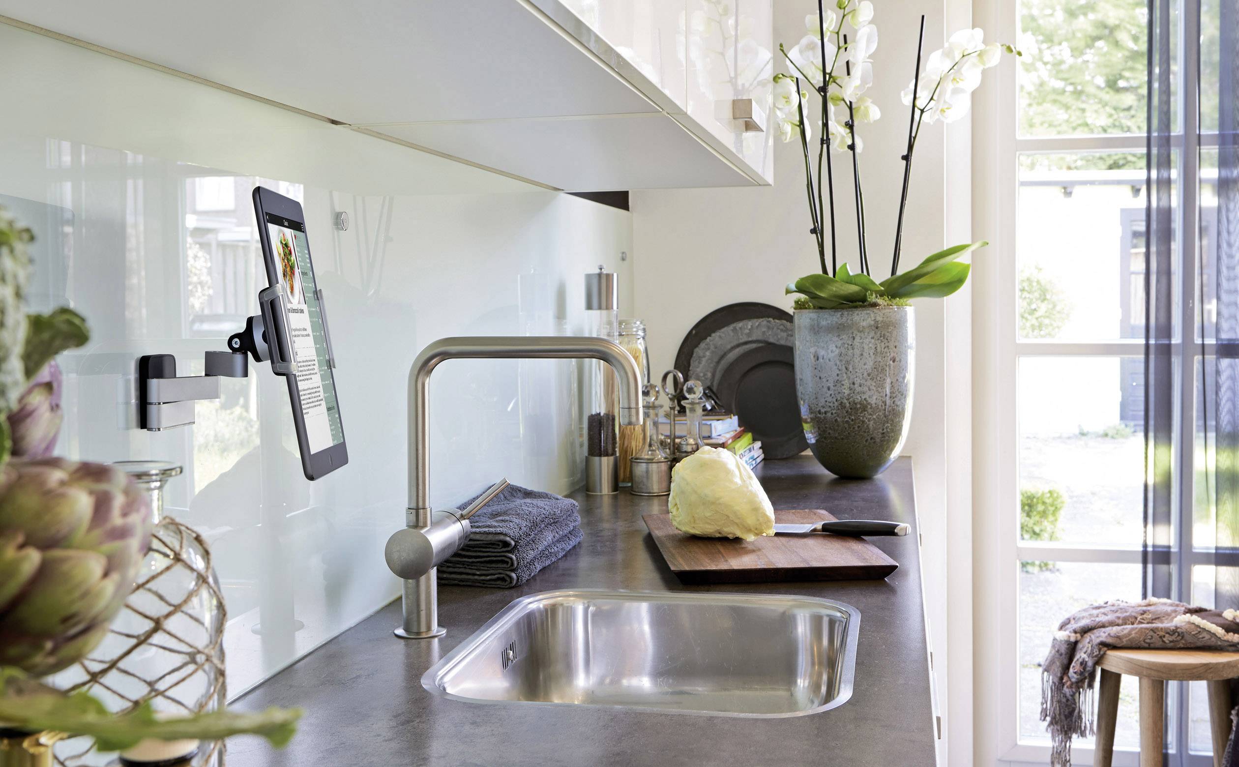 Moderne Küche mit Spüle, Schneidbrett und Tablet-Halterung. Eine Orchidee steht auf der Fensterbank. Helle, luftige Atmosphäre.