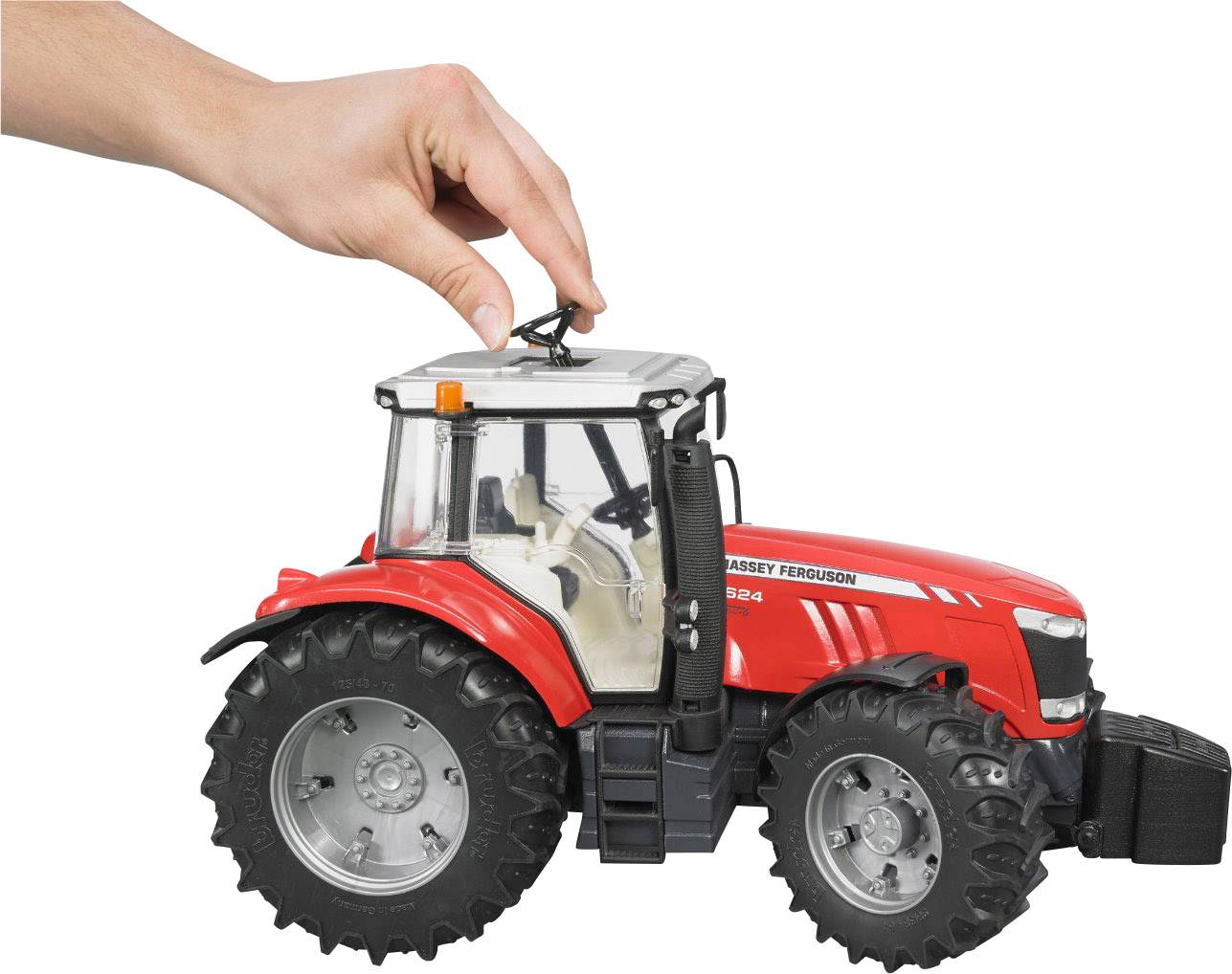 Eine Hand hält ein kleines, rotes Spielzeugtraktor-Modell mit großen Reifen, das auf einer weißen Oberfläche steht.