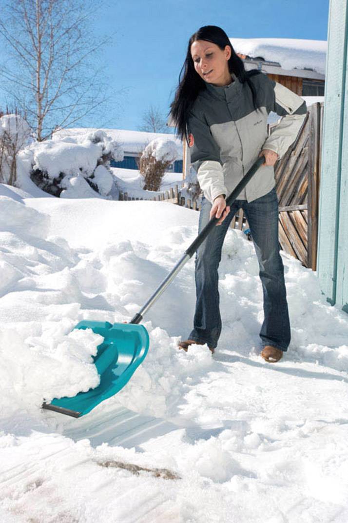 Frau schaufelt Schnee vor einem Haus, sonniger Wintertag, blauer Himmel im Hintergrund.