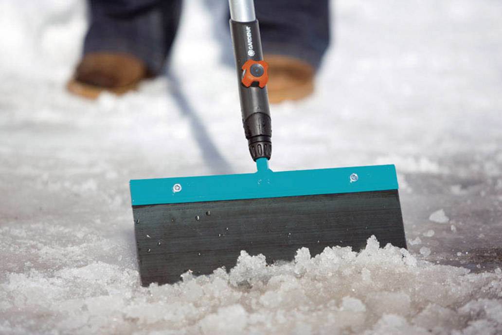 Person schiebt mit einem Schneeschieber Schnee von einem Gehweg. Im Vordergrund ist das blaue Schaufelblatt zu sehen.