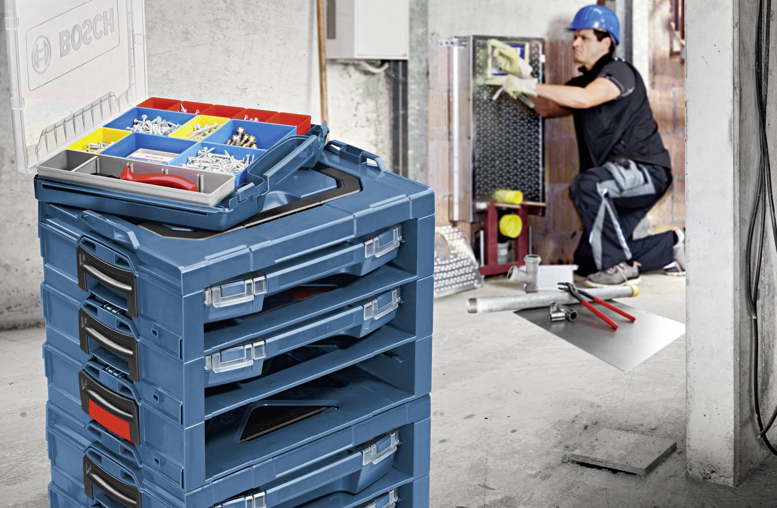 Ein Handwerker installiert an einer Wand Geräte. Im Vordergrund ist ein Stapel von blauen Werkzeugkästen mit sortierten Kleinteilen.