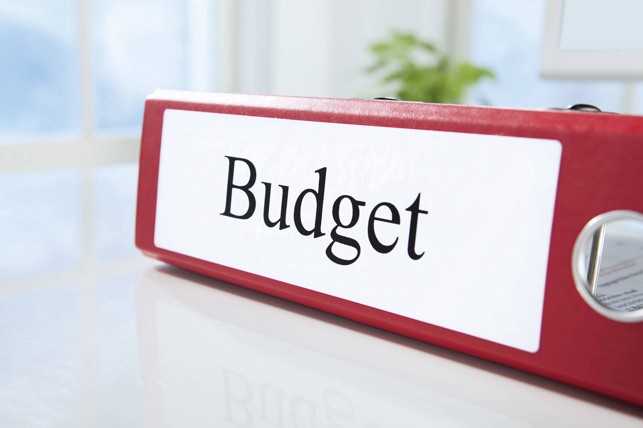 Roter Aktenordner mit der Aufschrift 'Budget' auf einem Schreibtisch im Büro, Hintergrund unscharf mit Fenster und Pflanzen.